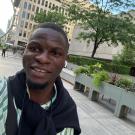 A selfie of a man in a green shirt and dark sweater standing in front of a building